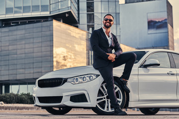Happy handsome bearded male in sunglasses dressed in a black suit sitting on luxury car against a...