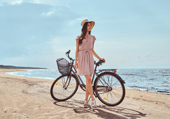 Beautiful brunette girl dressed in dress and hat posing with a bicycle on the beach on a sunny day.