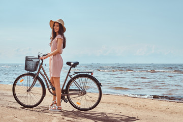 Fototapeta premium Beautiful brunette girl dressed in dress and hat posing with a bicycle on the beach on a sunny day.