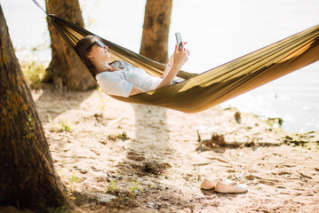Summer vacation concept, Happy young woman with sunglasses using a mobile phone in a hammock on the beach at sunset