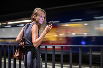 Pretty, young female commuter waiting for her daily train in a modern trainstation, using her cellphone while waiting (color toned image)