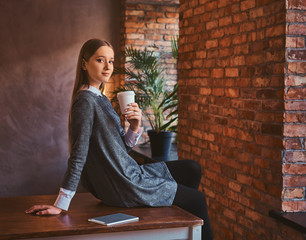 Fototapeta na wymiar Portrait of a young girl dressed in an elegant gray dress holds a cup of takeaway coffee looking at a camera while sitting on table in a room with loft interior.