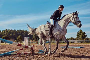 Female jockey on dapple gray horse jumping over hurdle in the open arena.