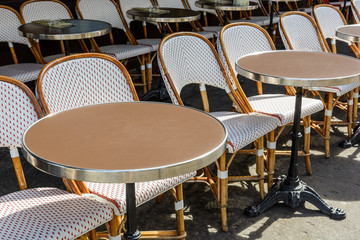 Round tables with a cast iron leg and rattan chairs lined up outside of a sidewalk cafe in Paris, France.