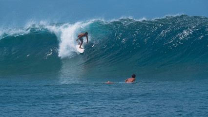 CLOSE UP: Young surfer watches his friend ride a huge emerald barrel ocean wave.