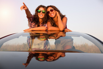 Two beautiful young girls in a convertible car looking at something at sunset