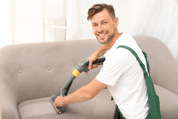 Male janitor removing dirt from sofa with upholstery cleaner indoors