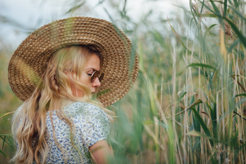 A blonde girl in a white dress with a blue print and a straw hat is walking among the reeds.