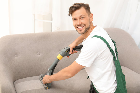 Male Janitor Removing Dirt From Sofa With Upholstery Cleaner Indoors