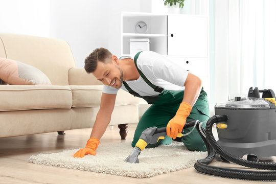 Male janitor removing dirt from rug with carpet cleaner in room