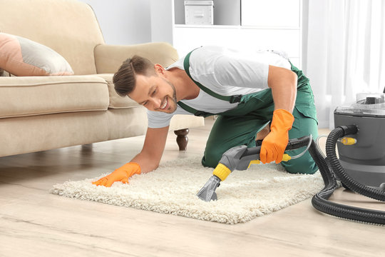 Male janitor removing dirt from rug with carpet cleaner in room