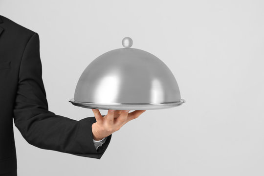 Waiter holding metal tray with lid on light background