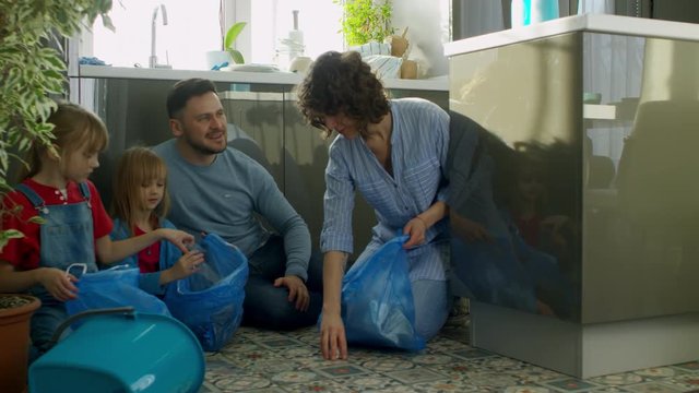 Family with two children of primary school age sitting on floor in kitchen, putting litter into different blue plastic bags and leaving room to throw it away