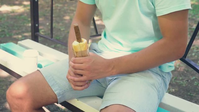 a young lonely guy holding a hotdog, in the lower abdomen, sitting on a bench in the park, and open the hotdog packaging, imitating an erection . 4k, slow-motion