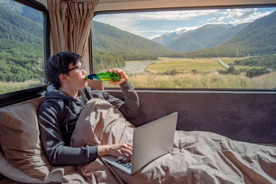 Young Asian man drinking beer and working with laptop computer on the bed in camper van with snow mountain scenic view through the window, digital nomad on road trip concept
