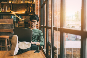 Asian man doing freelance work sitting out on a laptop computer connected to 4G Internet with a smart phone casual. By the window of a vintage coffee shop. - obrazy, fototapety, plakaty