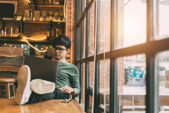 Asian man doing freelance work sitting out on a laptop computer connected to 4G Internet with a smart phone casual. By the window of a vintage coffee shop.