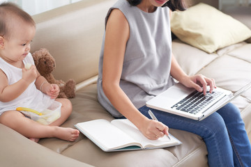 Obraz premium Young mother working on laptop and taking notes when her baby eating