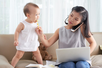 Freelance woman feeding her little baby when calling to client on phone