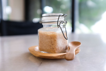 Close up of small glass jar of brown sugar on wooden tray with wooden spoon on a white marble table...
