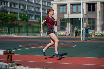 Fototapeta na wymiar Red-haired young girl runs in the stadium. Student delivers standards for running outdoors. Young woman running on the football field