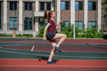 Red-haired young girl runs in the stadium. Student delivers standards for running outdoors. Young...