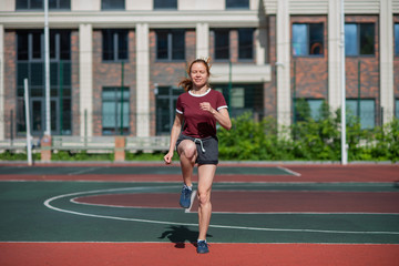 Fototapeta na wymiar Red-haired young girl runs in the stadium. Student delivers standards for running outdoors. Young woman running on the football field