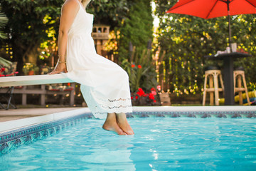 a woman in a white dress sitting on a diving board dipping her feet into a swimming pool
