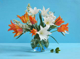 Bouquet of white and orange tulips and lilac in glass vase on blue background