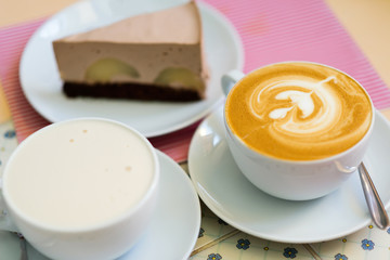 Сappuccino and latte (coffee with milk) on a pink-and white striped napkin with a chocolate cheesecake with apples in the background