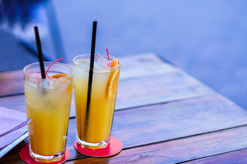 Two transparent high glasses of fresh orange beverage with drinking straws standing on red glass mats on wooden table in the evening outdoor.