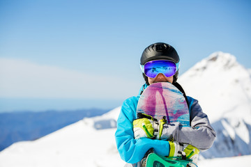 Fototapeta na wymiar Photo of young woman tourist in helmet looking into camera with snowboard in hands on background of snow mountains