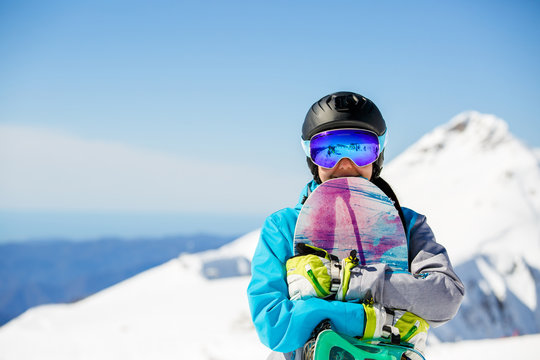 Photo of young woman tourist in helmet looking into camera with snowboard in hands on background of snow mountains
