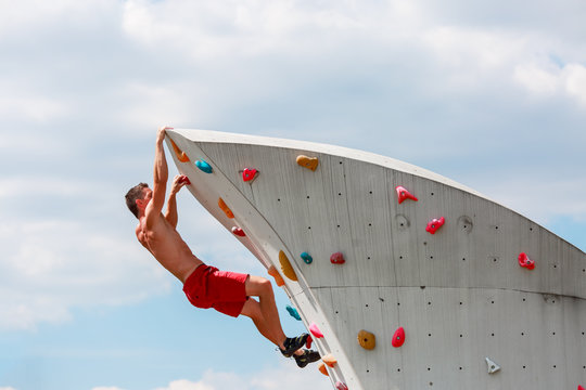 Photo of young sporty man in red shorts practicing on wall for rock climbing against blue sky with clouds