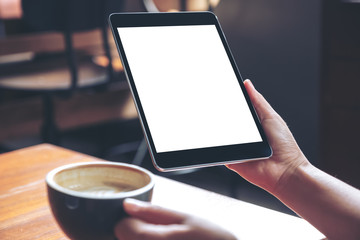 Mockup image of woman's hands holding black tablet pc with blank white screen with coffee cup on wooden table in cafe