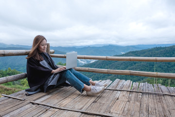 Closeup image of a woman working and typing on laptop while sitting on wooden balcony with green mountains on foggy day with blue sky background