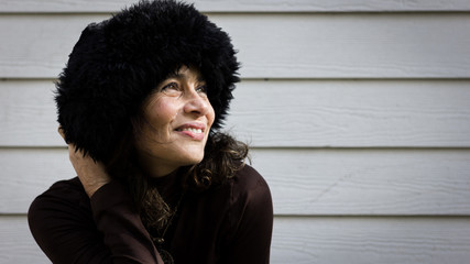 Fototapeta premium Smiling mature lady holds black fur Russian hat on head over white wood plank wall background. Middle age pretty woman happy on cold weather. Ready for winter season. Female fashion, trendy concepts