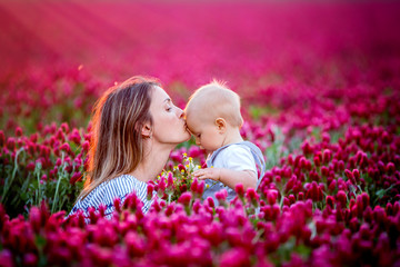 Young mother, embracing with tenderness and care her toddler baby boy in crimson clover field, smiling happily