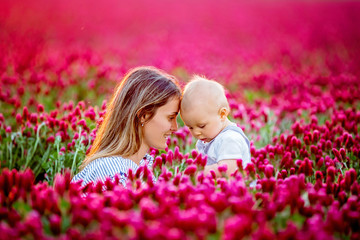 Young mother, embracing with tenderness and care her toddler baby boy in crimson clover field, smiling happily