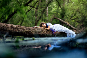 photo shoot of a girl with dreadlocks in a white dress in the nature near the water. the girl lies on the tree