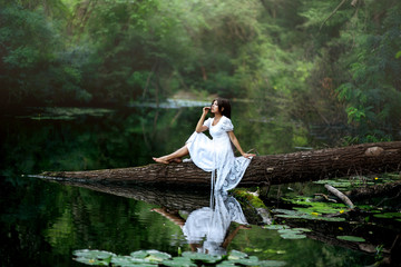 photo shoot of a girl with dreadlocks in a white dress in the nature near the water. the girl sits on a tree
