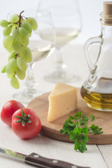 On the table is cheese, tomatoes, grapes, olive oil, parsley, two wineglasses and a bottle of white wine. Also on the white table is a cutting board and a knife.
