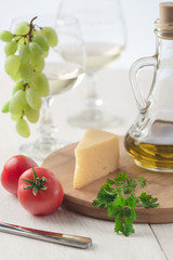 On the table is cheese, tomatoes, grapes, olive oil, parsley, two wineglasses and a bottle of white wine. Also on the white table is a cutting board and a knife.