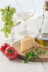 On the table is cheese, tomatoes, grapes, olive oil, parsley, two wineglasses and a bottle of white wine. Also on the white table is a cutting board and a knife.