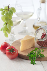 On the table is cheese, tomatoes, grapes, olive oil, parsley, two wineglasses and a bottle of white wine. Also on the white table is a cutting board and a knife.