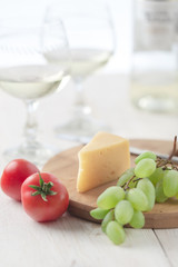On the table is cheese, tomatoes, grapes, olive oil, parsley, two wineglasses and a bottle of white wine. Also on the white table is a cutting board and a knife.