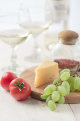 On the table is cheese, tomatoes, grapes, olive oil, parsley, two wineglasses and a bottle of white wine. Also on the white table is a cutting board and a knife.