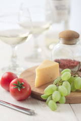 On the table is cheese, tomates, grapes, olive oil, parsley, two wineglasses and a bottle of wine.