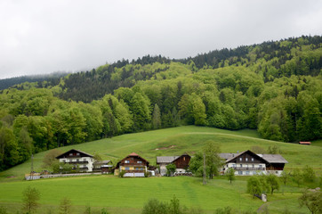 Fototapeta na wymiar Some typical Austrian houses, three stories in height with wooden sloping roofs, are in the foot of the alps. The hills behind are lush and green, and covered in forest The sky is overcast.