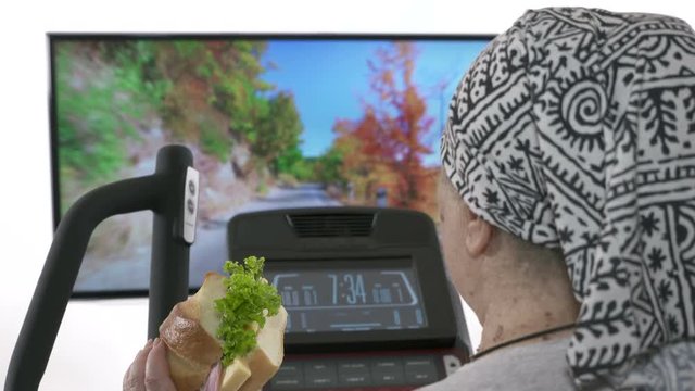 Obese Senior Person Eating Fatty Sandwich During Exercise On Elliptical Trainer